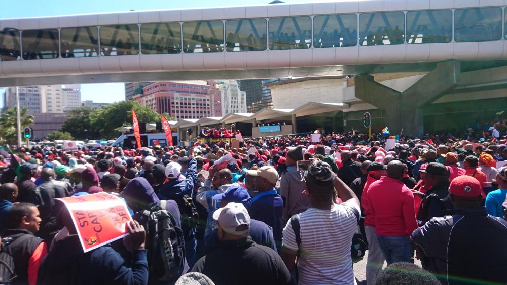 Color photo of many protestors facing away from the camera, one protestor holding a sign covering their face. Above the protestors, there is a see-through bridge and behind that, a city landscape. 