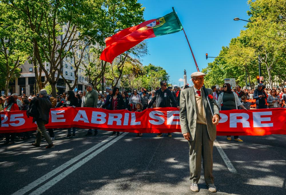 A man in a grey suit stands in the street holding a Portugese flag waving; behind him, demonstrators carry a red banner with white writing.